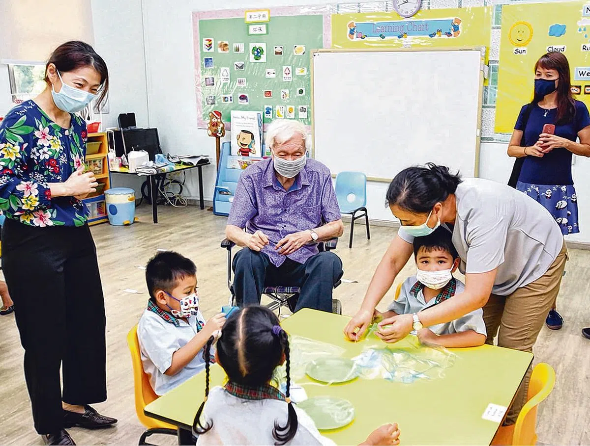 星洲幼稚园在百年大庆纪念年邀请创校人郑聘廷牧师的后人到访，参观幼童学习美工劳作，坐轮椅者是郑庆岩，右一是郑妙菁，左一是幼儿园校长萧慧敏。（陈福洲摄）