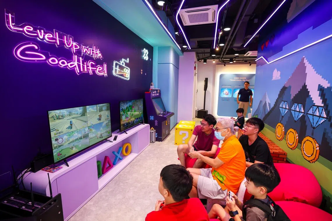 位于淡滨尼第142座组屋底层的乐龄中心Goodlife Studio主打亲家庭活动，让年长居民与年轻一辈拉近关系。洪昭瑜（身着橙色衣）常到中心活动，在儿子洪毅铭（身着红衣）的引导下，尝试电玩游戏。（林明顺摄）