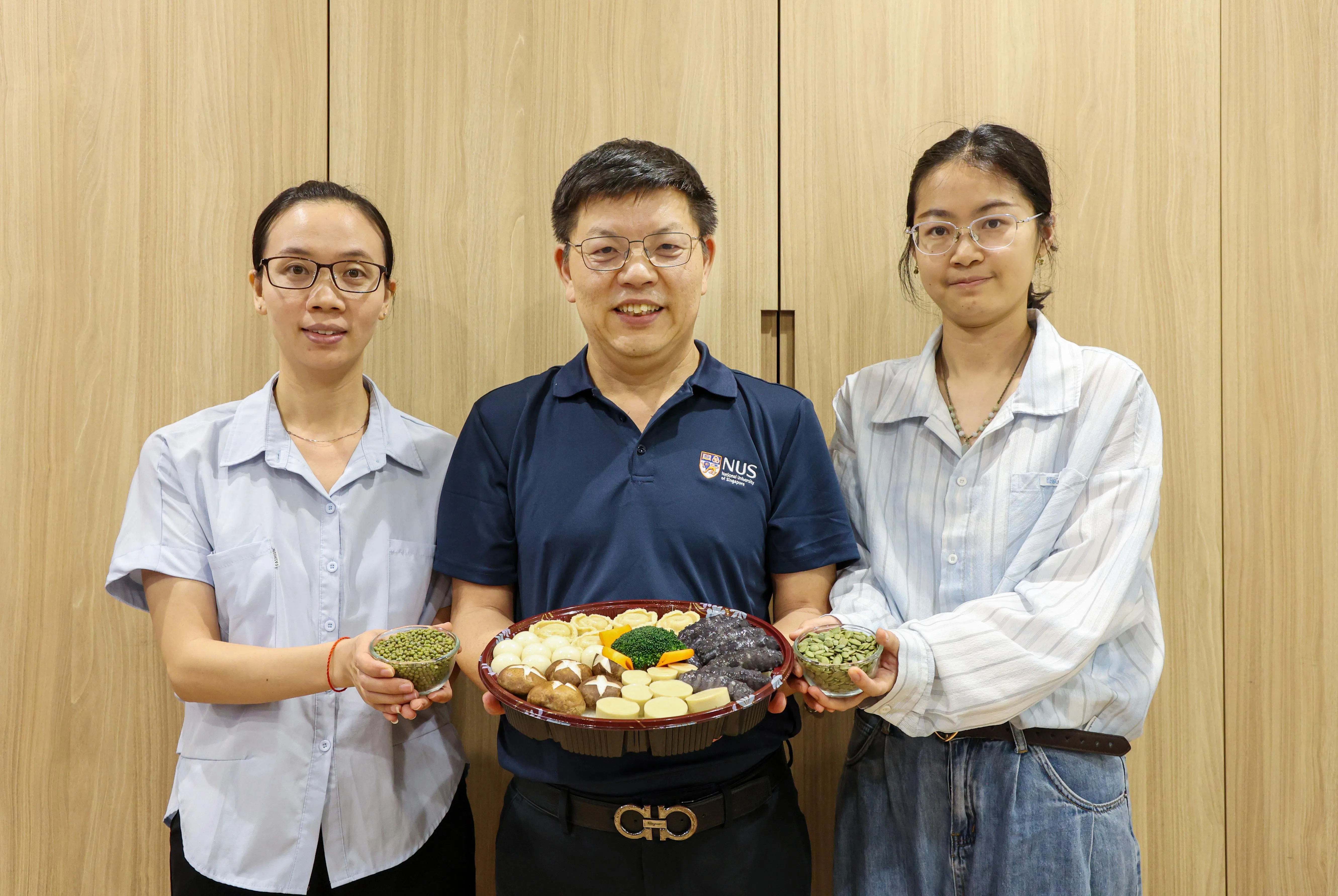 食品科学与工程系副主任黄德建教授（中）手捧的盆菜暗藏玄机，其中鲍鱼由绿豆制成，海参则源自南瓜籽。这项创新由研究员刘云娇博士（左，手持绿豆）与林祎博士（右，手持南瓜籽）共同参与研发。（游甯稀摄）