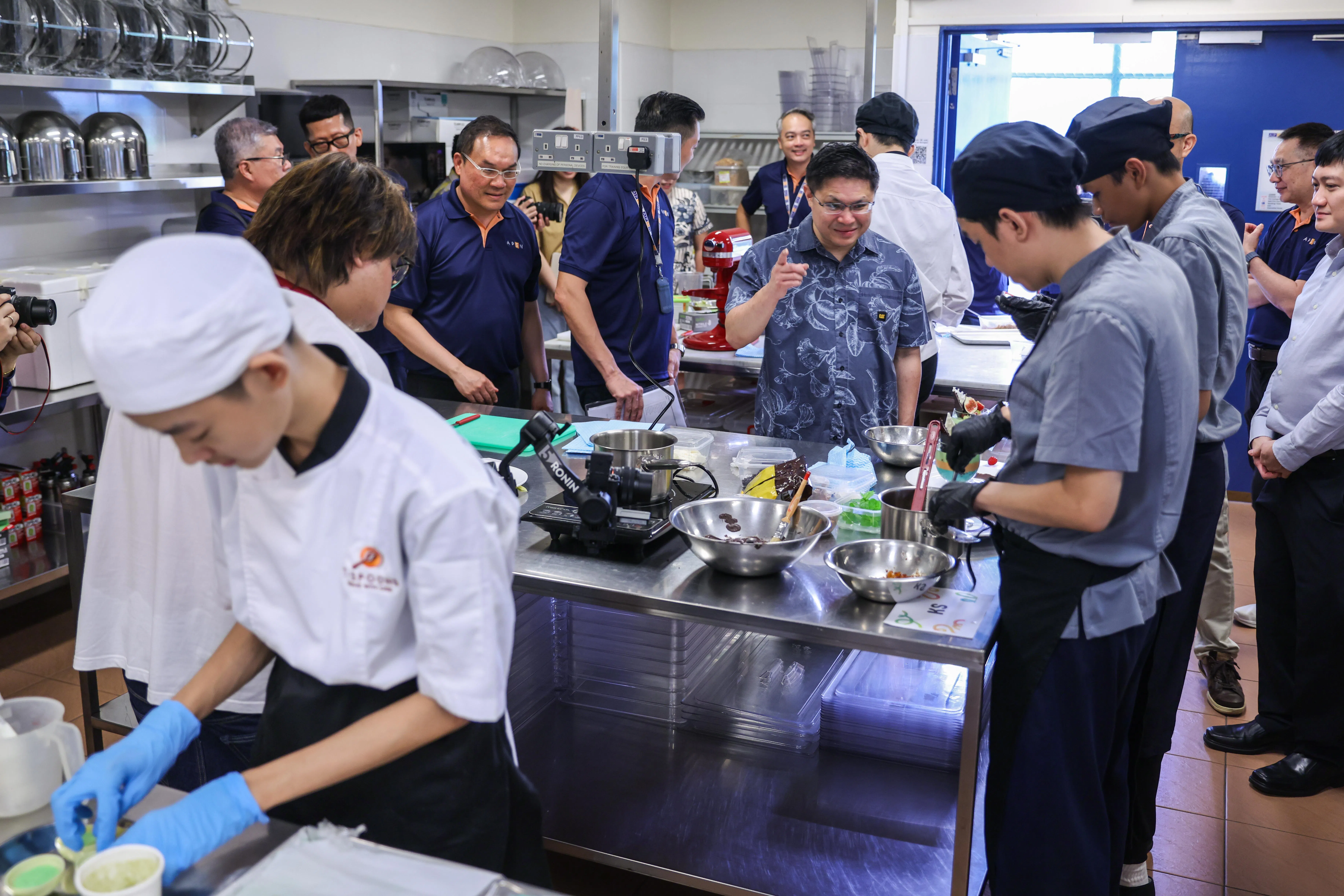 人力部兼能源贸工部政务部长符策翔（蓝色花纹上衣）参观“ONE APSN技能挑战赛展示会2026”的烹饪与餐饮区，观看特需学员在专业厨房制作料理，了解他们所掌握的烹饪技能和工作习惯。（白艳琳摄）