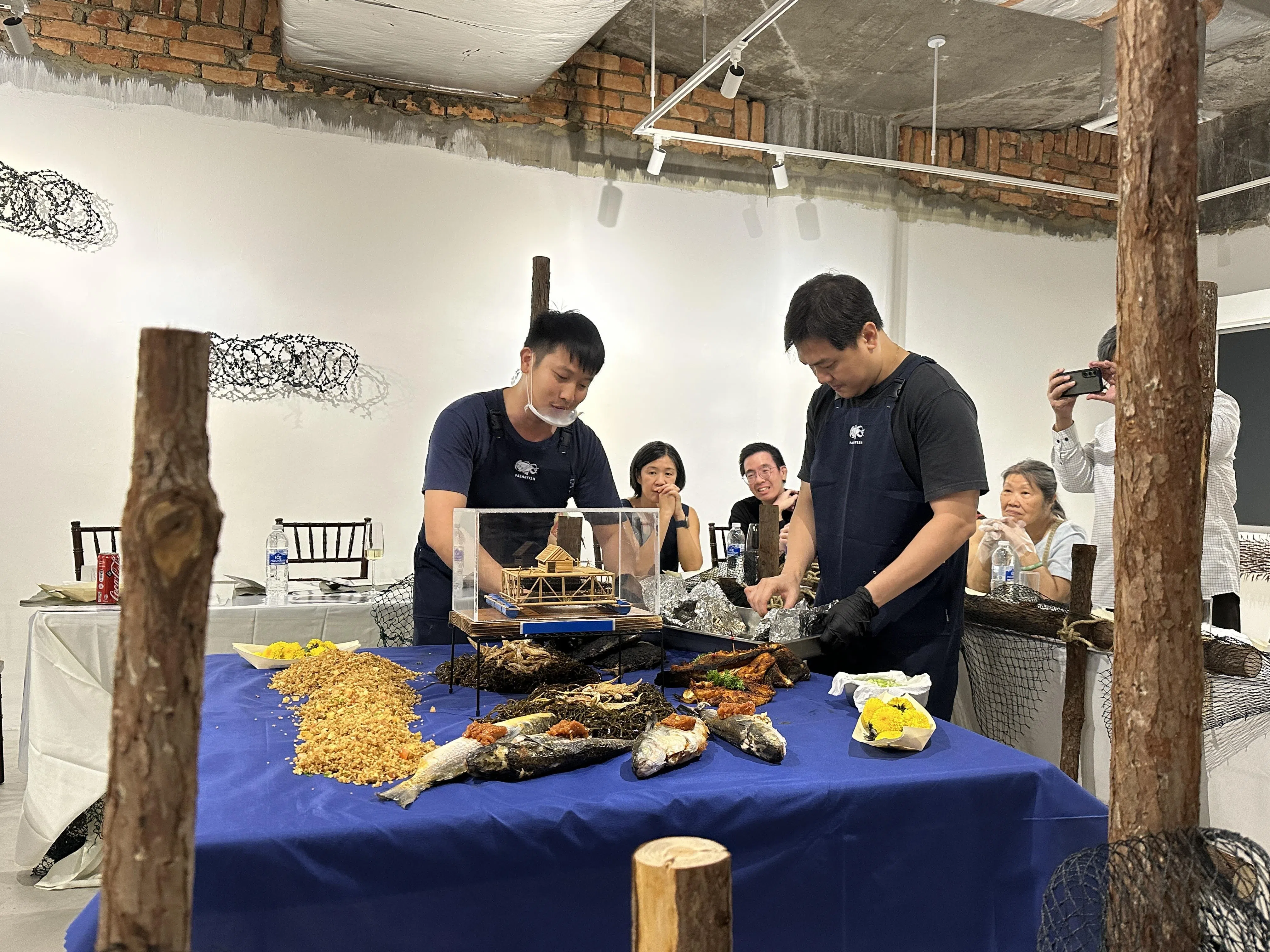 “巴刹鱼”联合创办人陆勇义（站者左一）在布置海鲜晩餐。桌子周围与墙面为艺术家庄志伟（坐者左二）的装置作品，E63奎笼主人黄梅莲（坐者右一）坐在一旁。（黄向京摄）